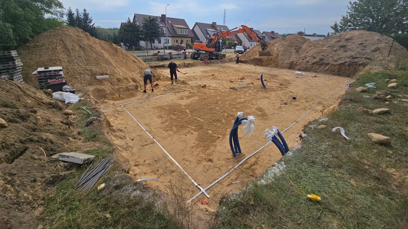Piaskowa podbudowa pod płytę fundamentową z ułożonym uziomem i przepustami na kable, w tle koparka