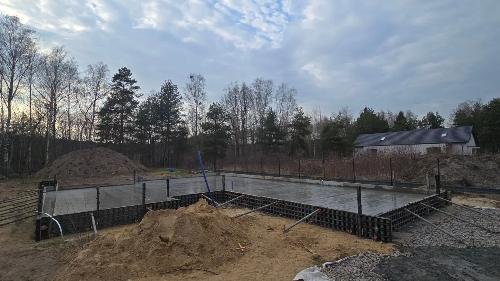 Płyta fundamentowa po betonowaniu w szalunku systemowym, w tle las i dom