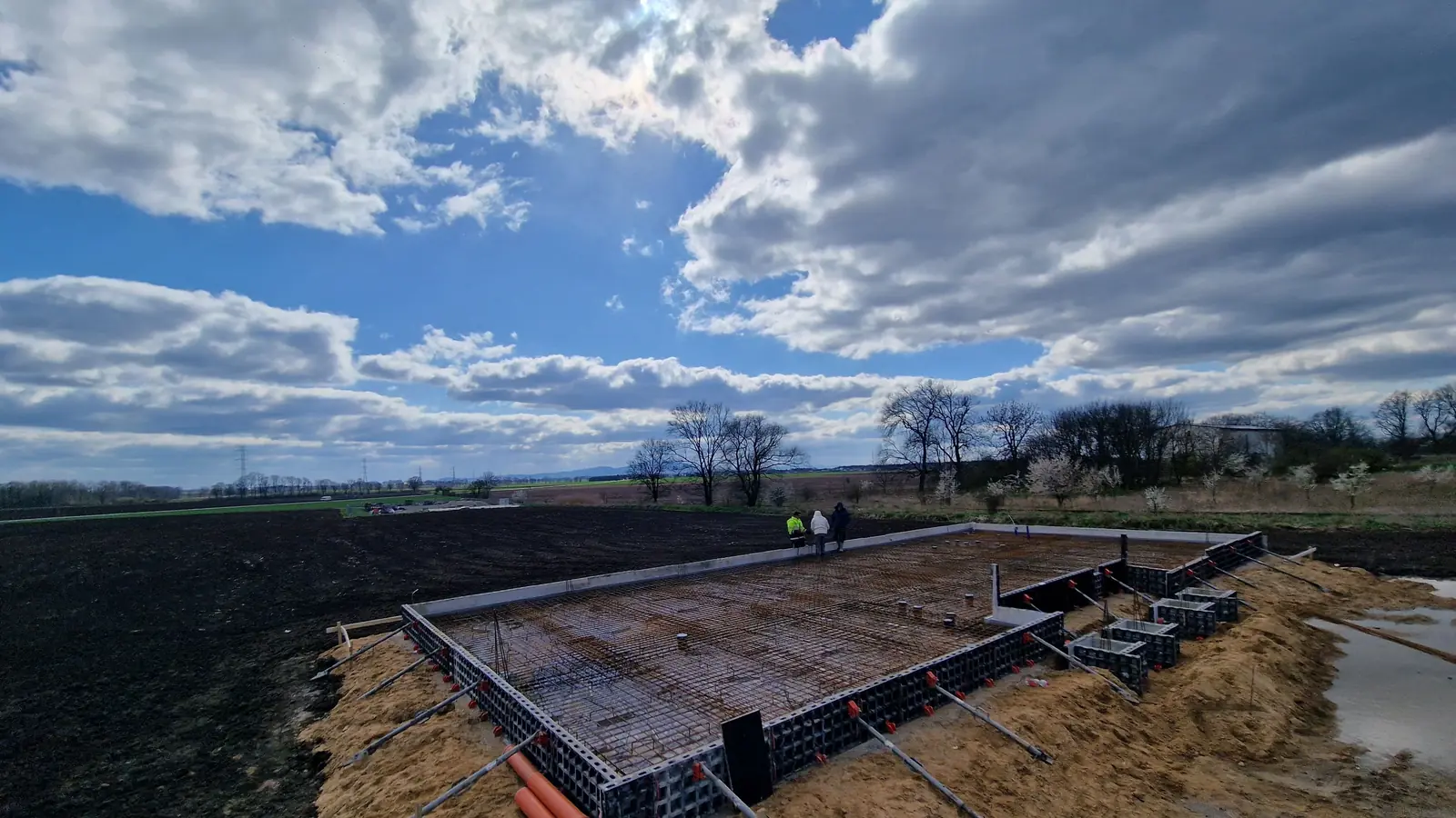 Zbrojenie płyty fundamentowej w szalunku systemowym na otwartej działce, nad nią zachmurzone niebo